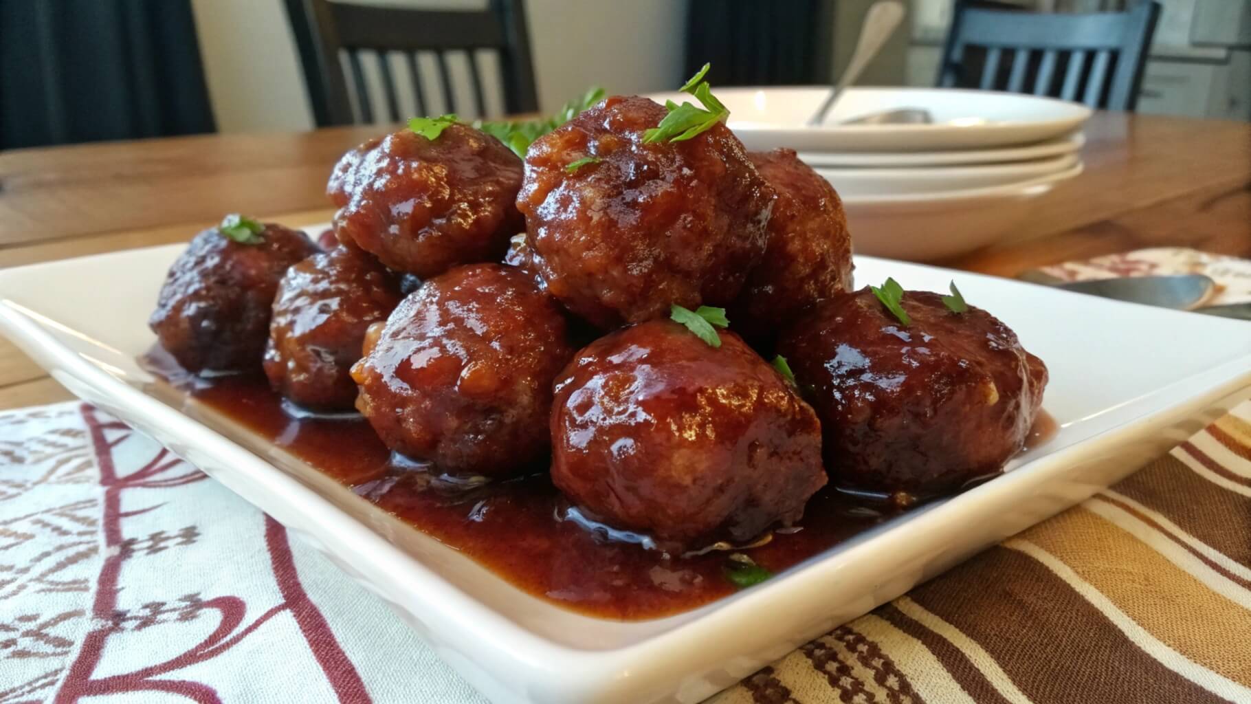 A plate of glazed meatballs stacked on a white dish, sitting on a red-and-white checkered cloth. The meatballs are coated in a rich, glossy brown sauce, with a serving dish of more meatballs in the background on a wooden table in a kitchen setting.