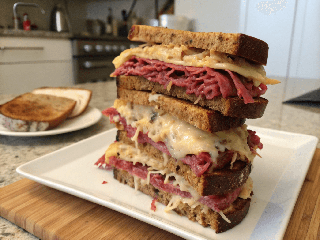 Is a Reuben sandwich made with pastrami? This towering sandwich features layers of corned beef, melted cheese, and sauerkraut on toasted rye bread in a kitchen setting.